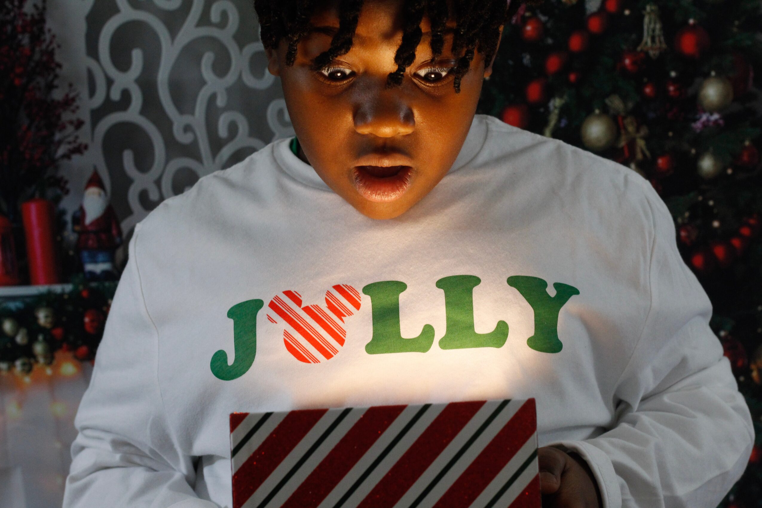 Child with surprised expression wearing a white shirt with holiday-themed text, in front of a Christmas tree.