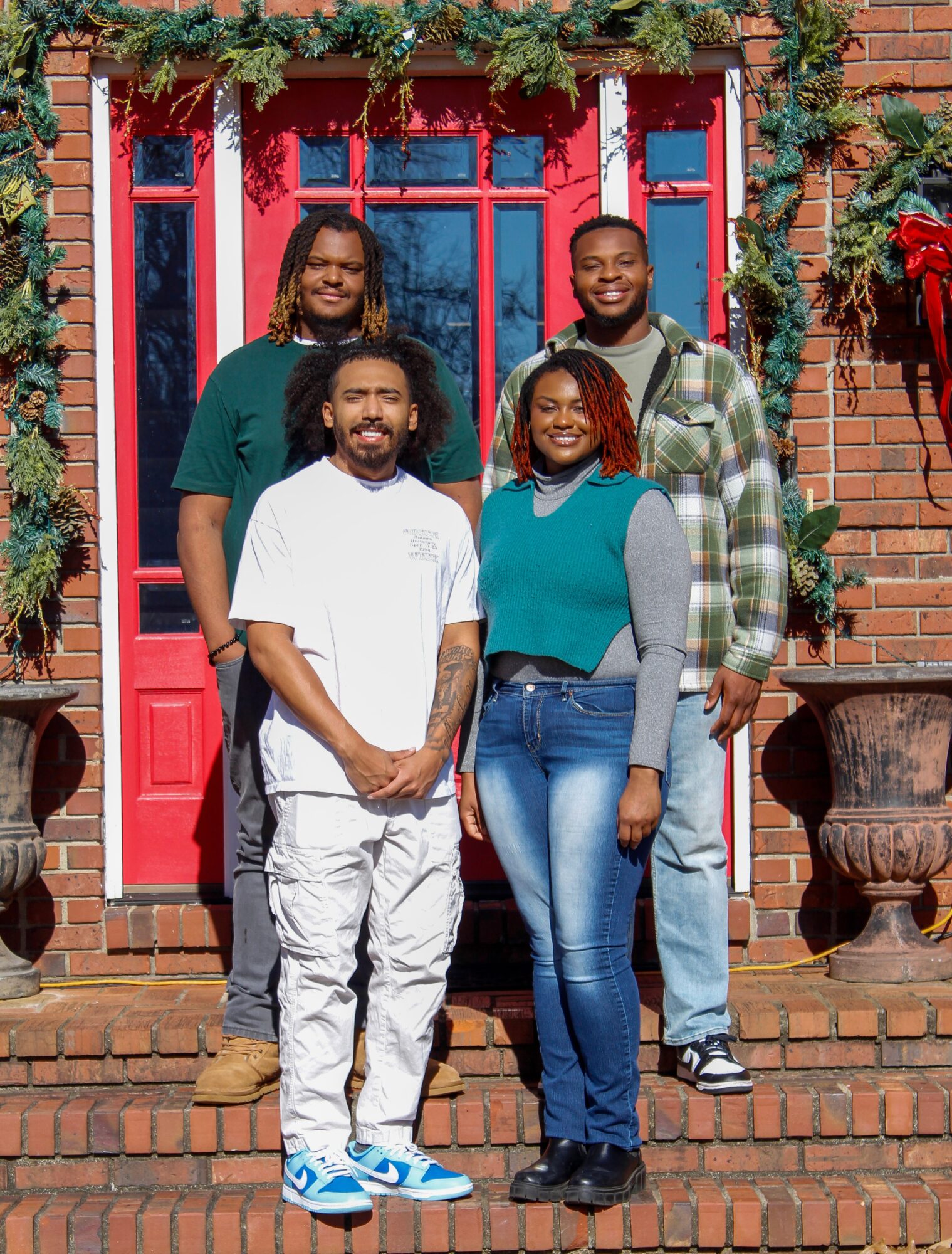 Five people stand on steps in front of a red door with greenery and brick wall, smiling at the camera.