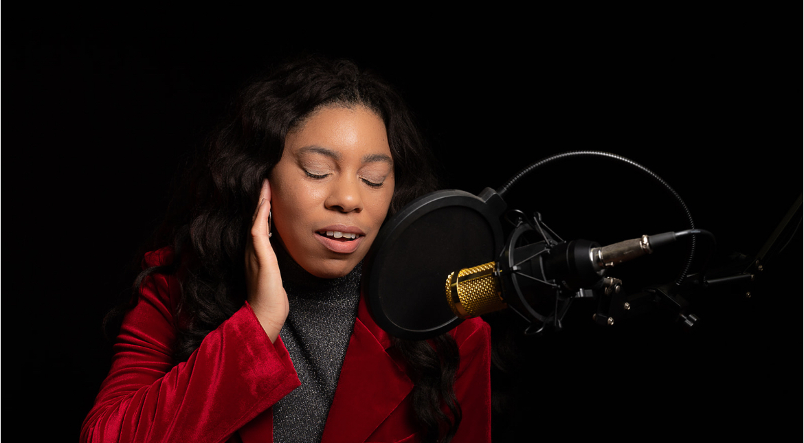 Woman singing into a microphone with eyes closed, wearing a red jacket and gray top, against a black background.