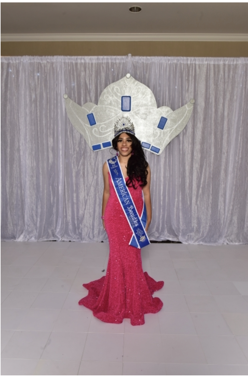 Woman in pink gown with crown and sash standing in front of silver curtain backdrop.