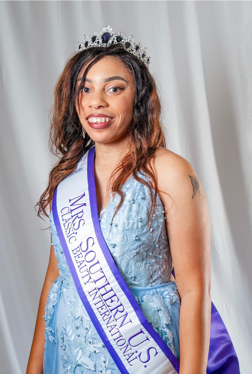 Young woman wearing a crown and sash, smiling, in front of a white curtain, dressed in a light blue gown.
