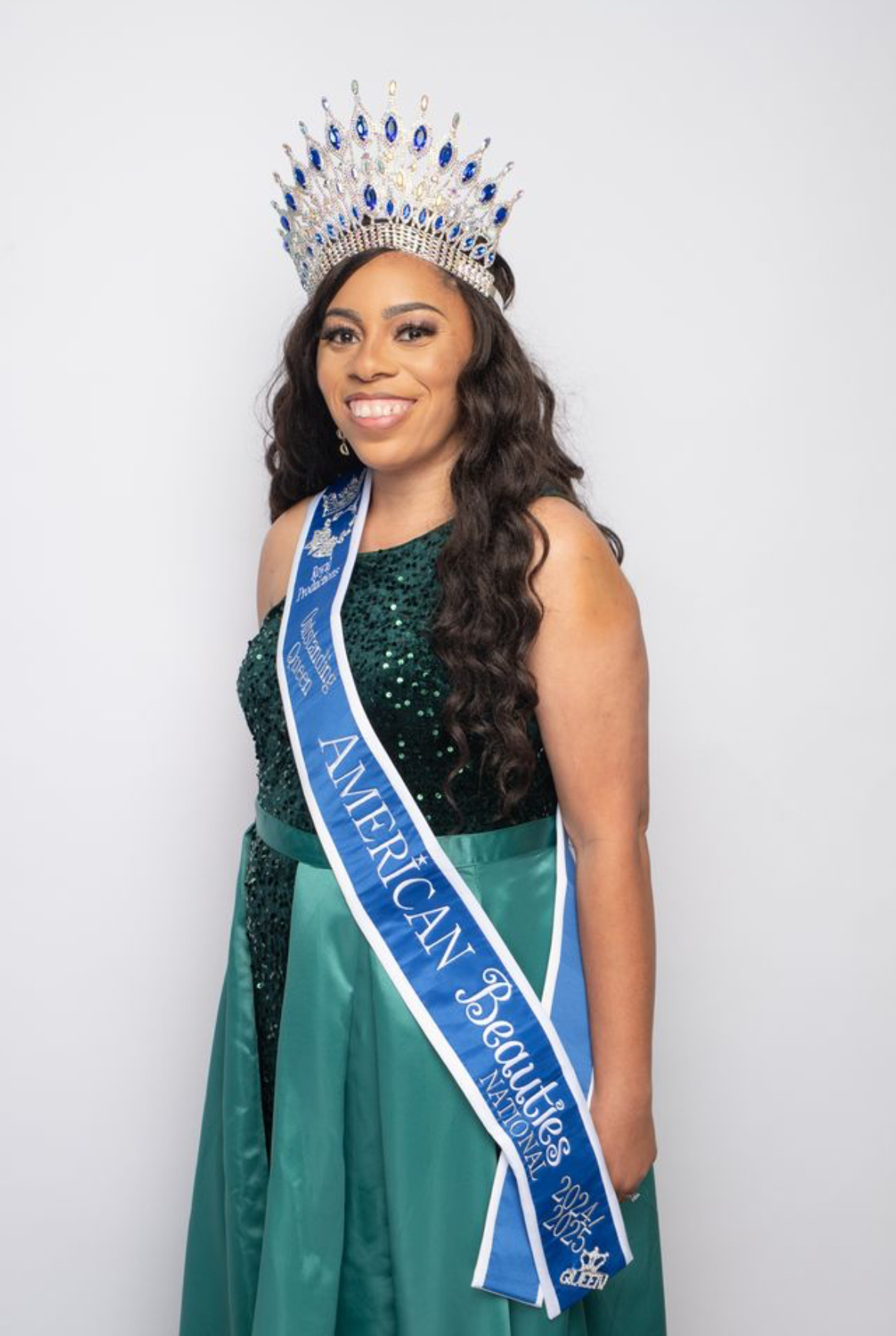 Woman wearing a crown and sash, smiling, in a green dress, standing against a plain background.