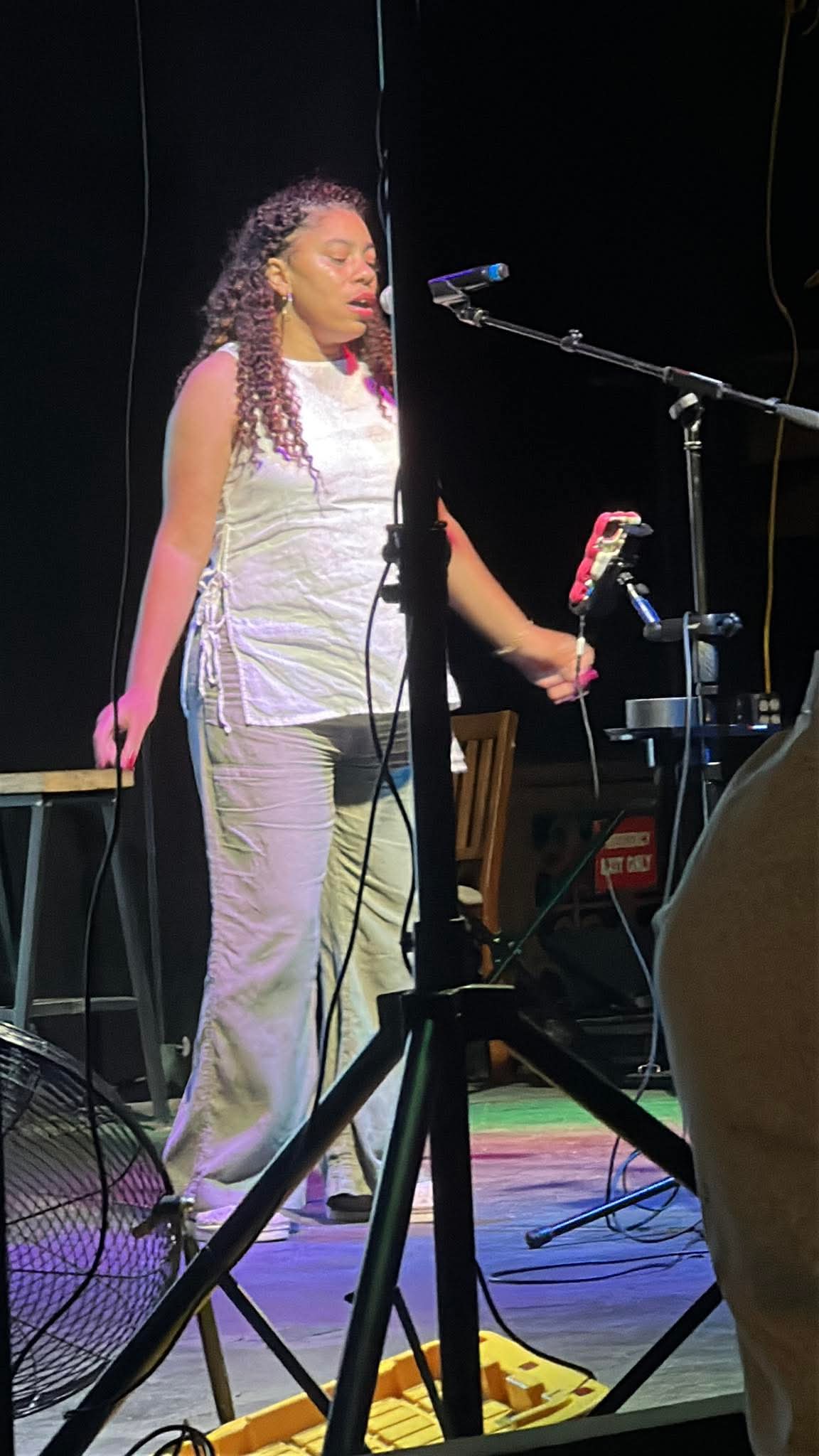 Woman standing on stage with microphone, holding a phone, surrounded by musical equipment, in a dark setting.