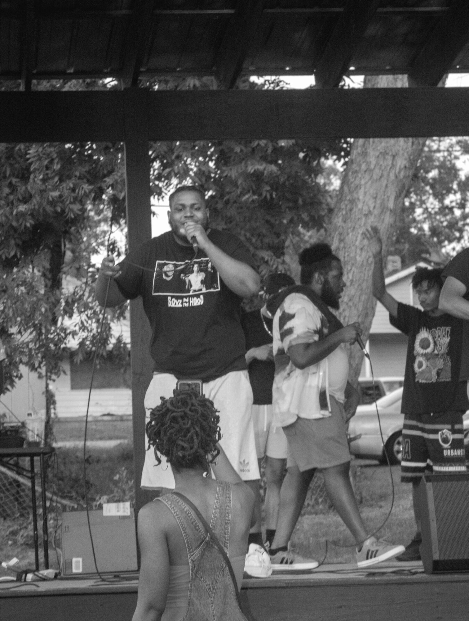 Group of people performing on an outdoor stage with trees in background, one person singing into a microphone, others dancing.
