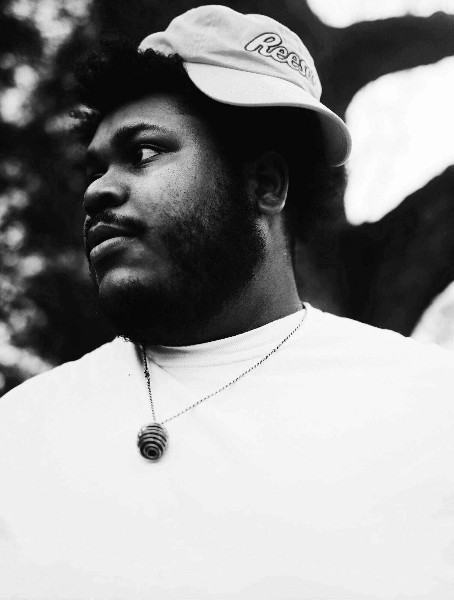 Young man with a hat and necklace, looking to the side outdoors, black and white photo.