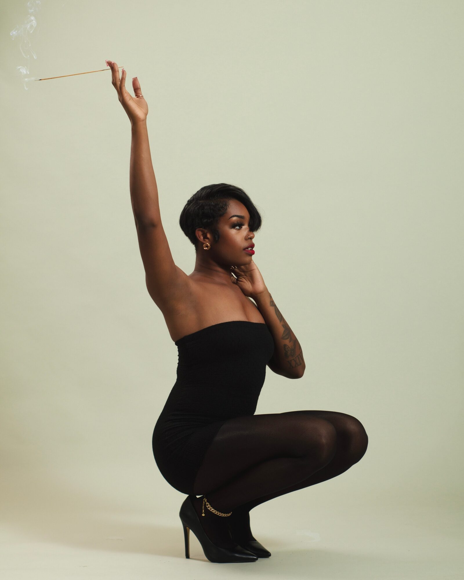 Woman in black dress and tights squats with one arm raised holding a stick, against a plain light background.