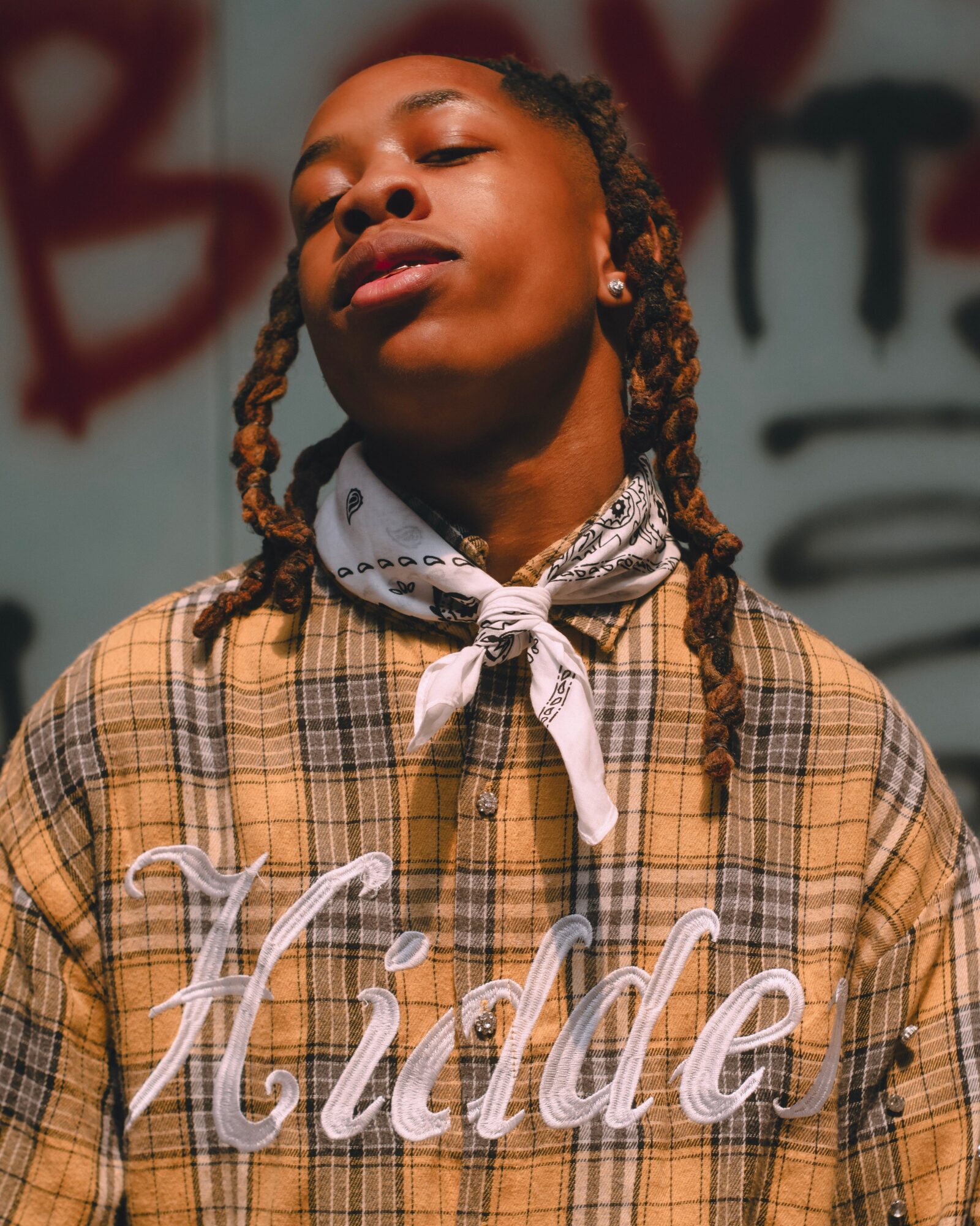 Young person with braided hair wearing a plaid shirt and bandana, standing in front of graffiti background.