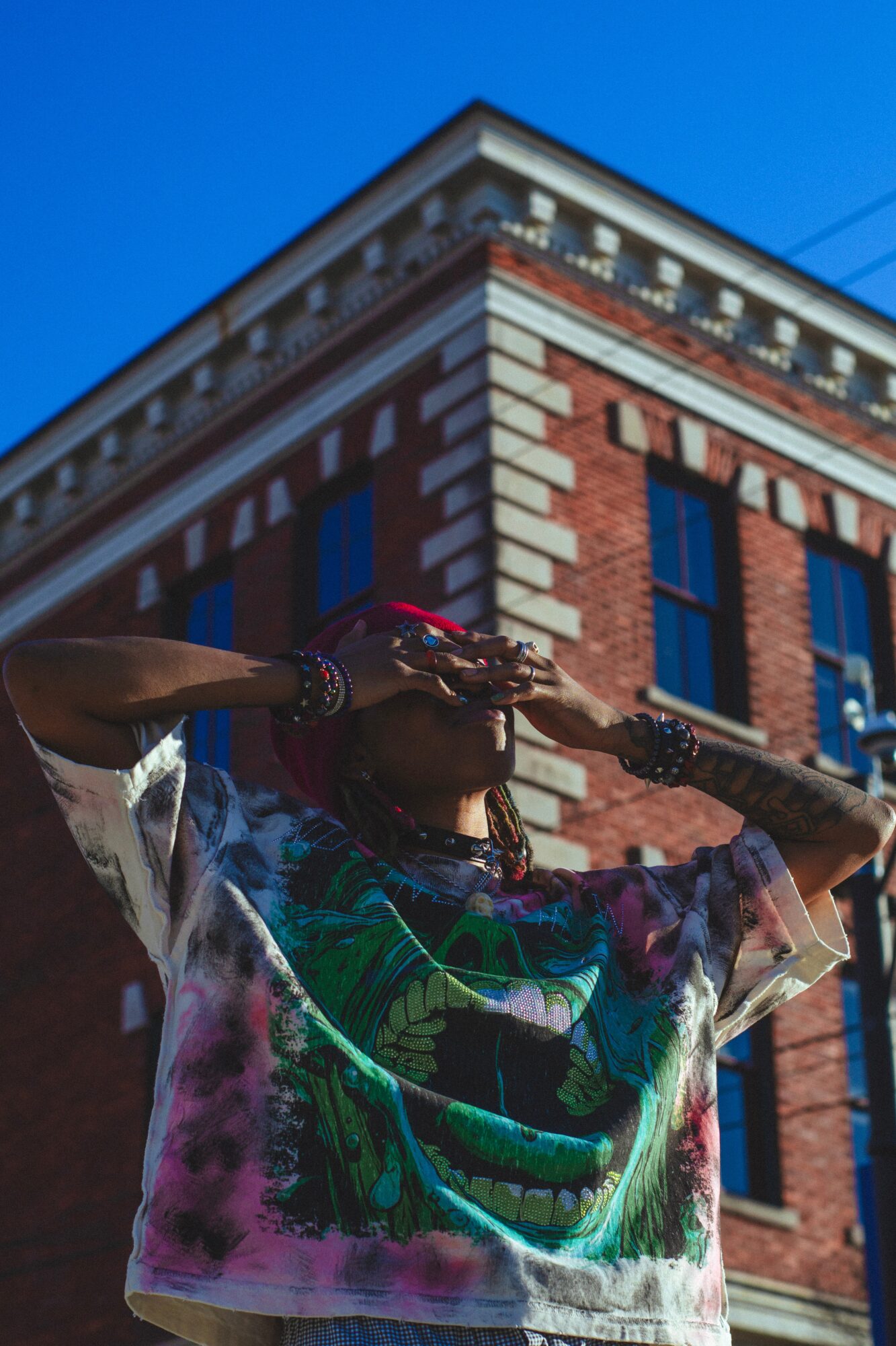 Person with colorful clothing and jewelry standing in front of a brick building under a blue sky.