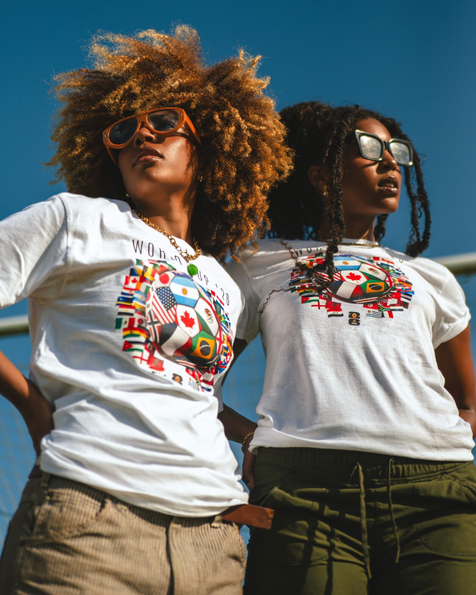 Two women with curly hair wearing sunglasses and white T-shirts with colorful designs, standing outdoors against a blue sky.