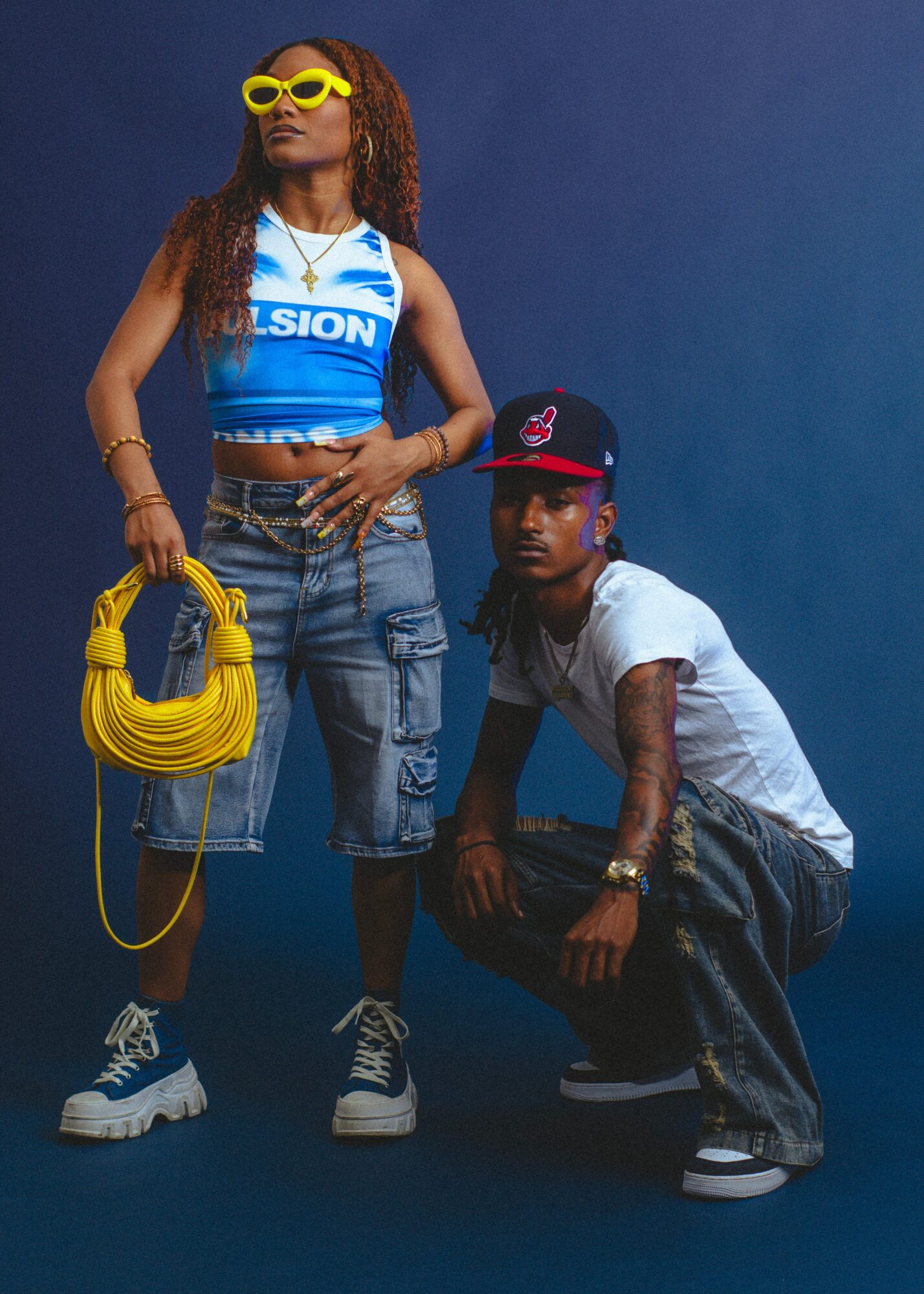 Two young people pose against a dark blue background, one standing with yellow sunglasses and a yellow bag, the other crouching.