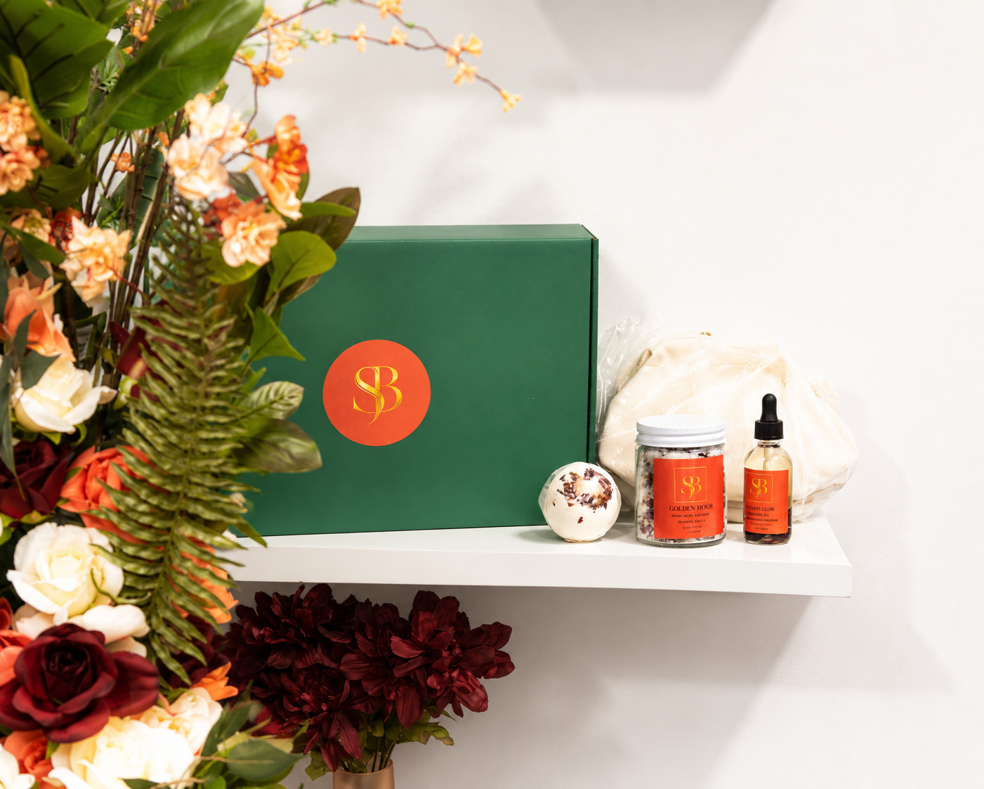 White shelf with a green box, candles, and a dropper bottle, next to a large floral arrangement, against a white wall.
