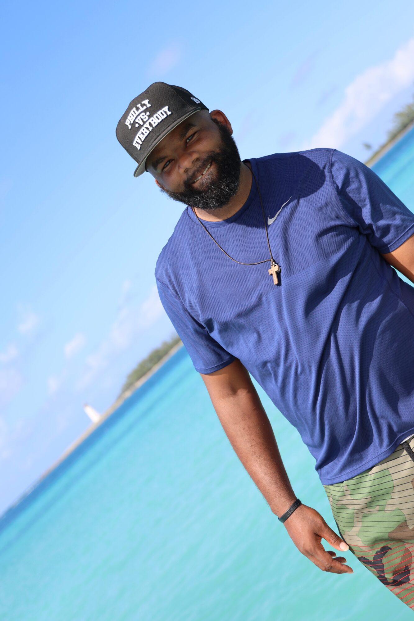 Man with beard wearing a black cap, blue shirt, and sunglasses stands near water with a blue sky background.