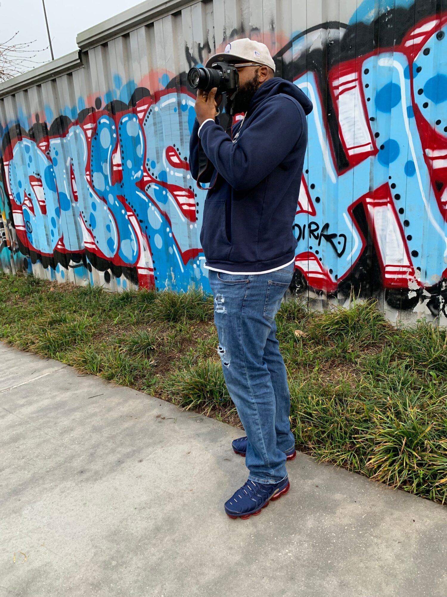 Person with a backpack and white cap standing next to a graffiti-covered wall, sidewalk, and grass.