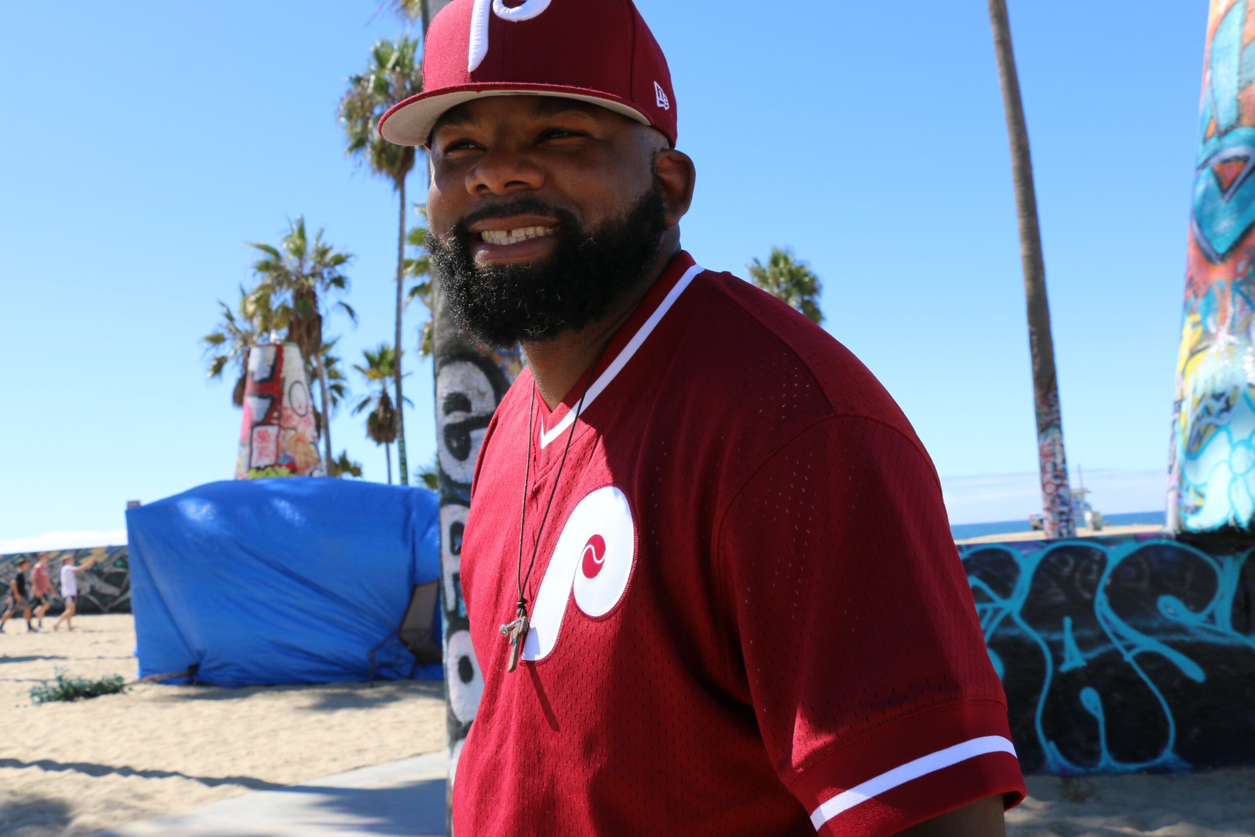 Man with a beard wearing a maroon Philadelphia Phillies jersey and cap, smiling outdoors on a sunny day.