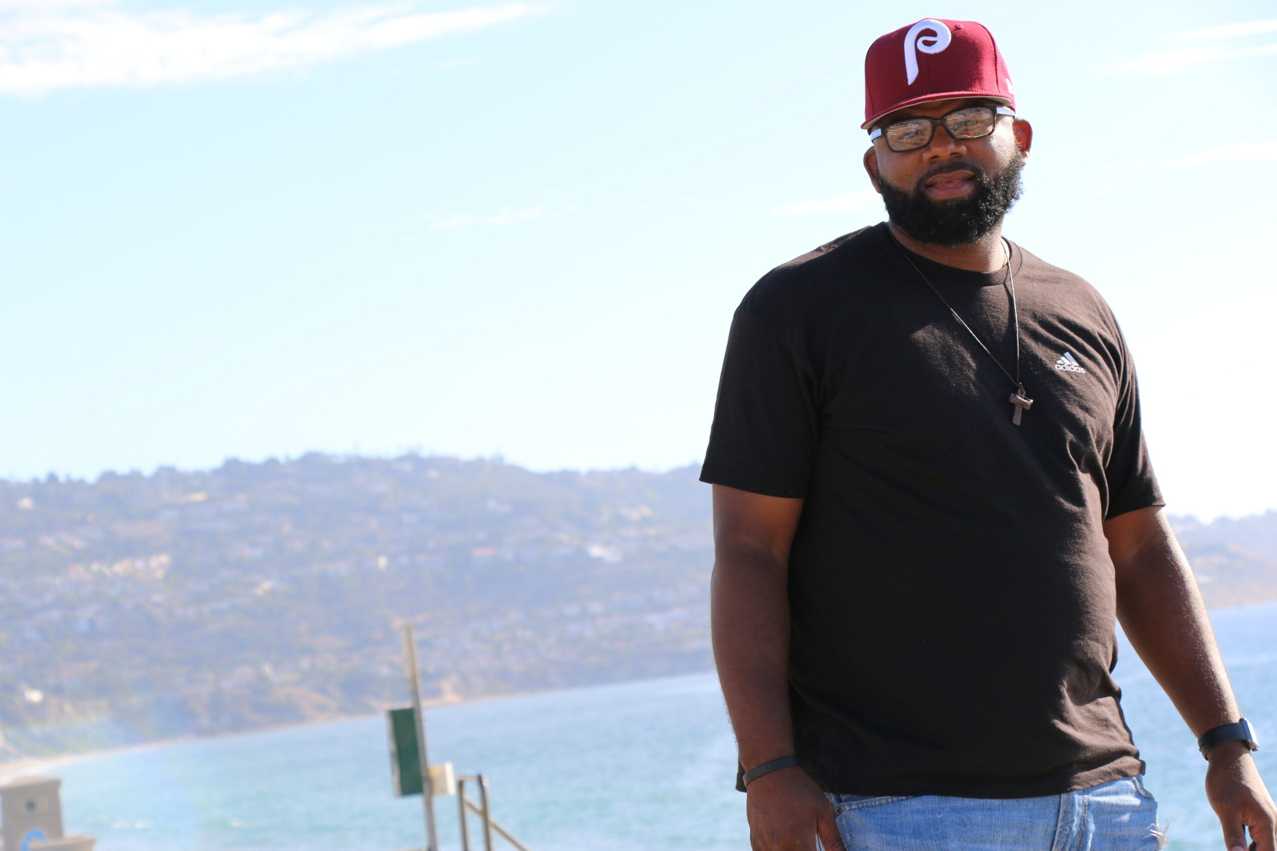 Man with glasses, beard, black t-shirt, red cap, standing outdoors near water, with hills in background.