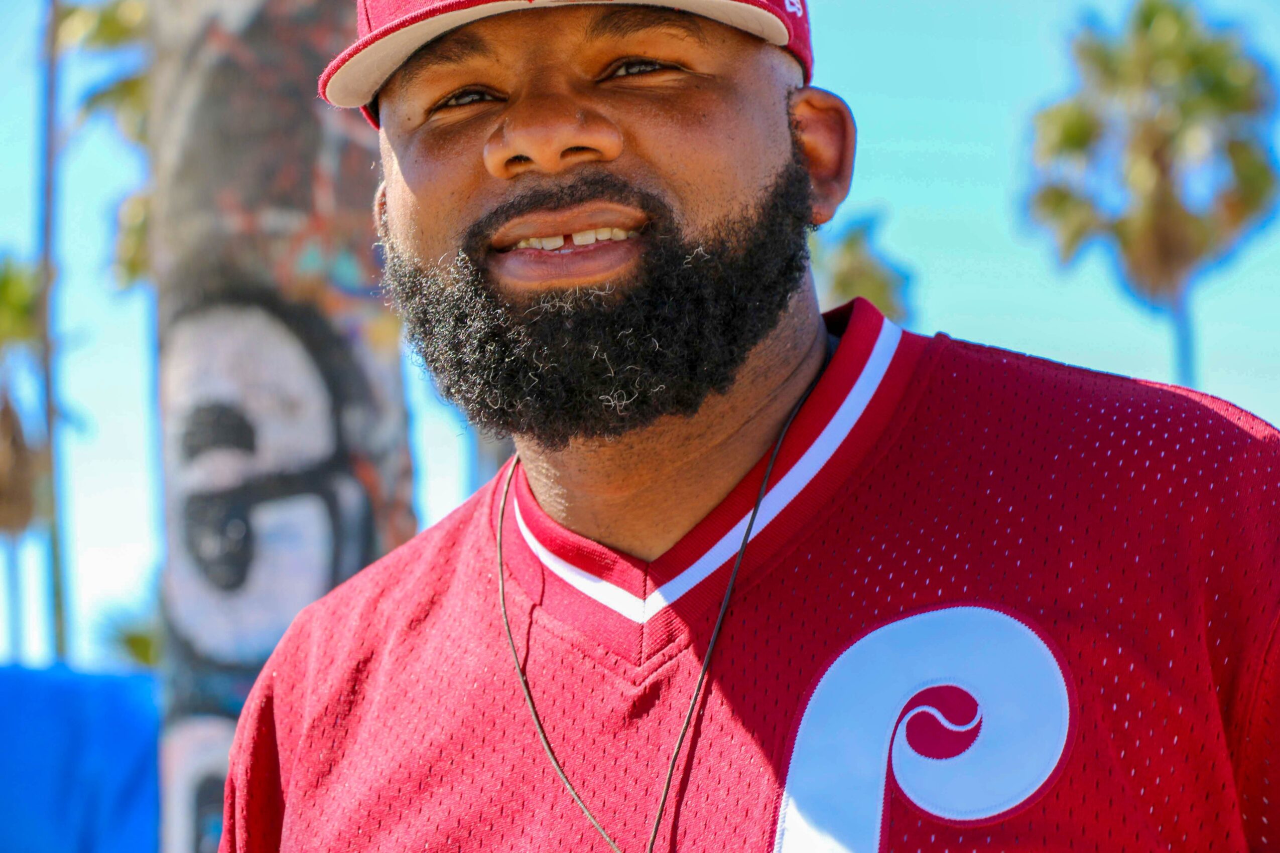 Close-up of a man with a beard wearing a red sports jersey and a cap outdoors, with palm trees in the background.