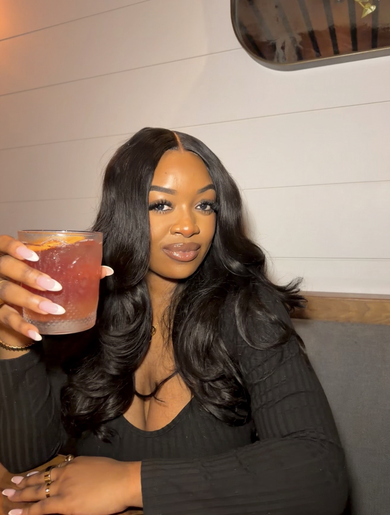 Woman with long wavy hair holding a drink, sitting indoors, smiling at the camera.