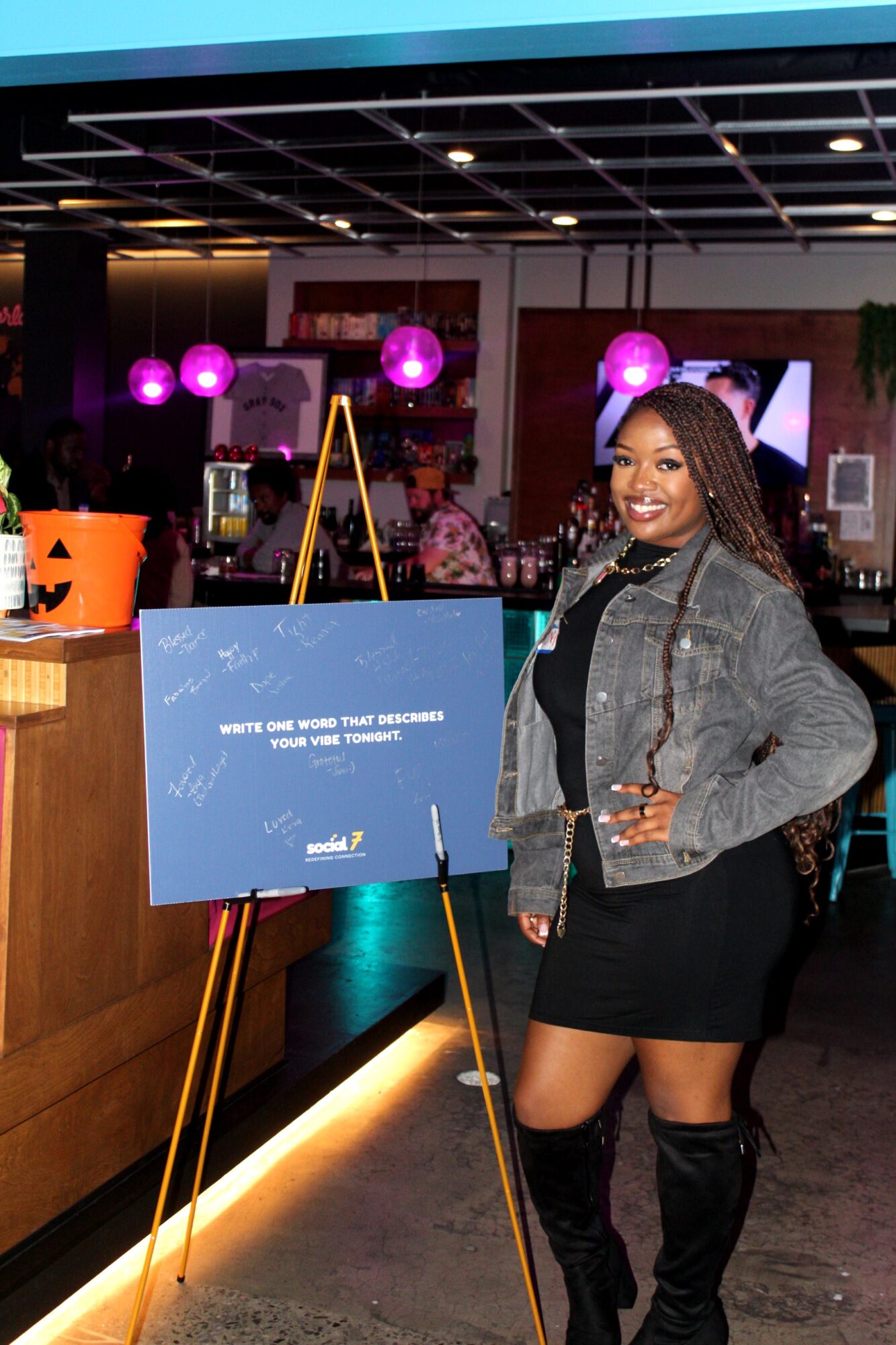 Woman smiling in a gray jacket and black dress standing next to a blue sign on an easel in a decorated indoor space.