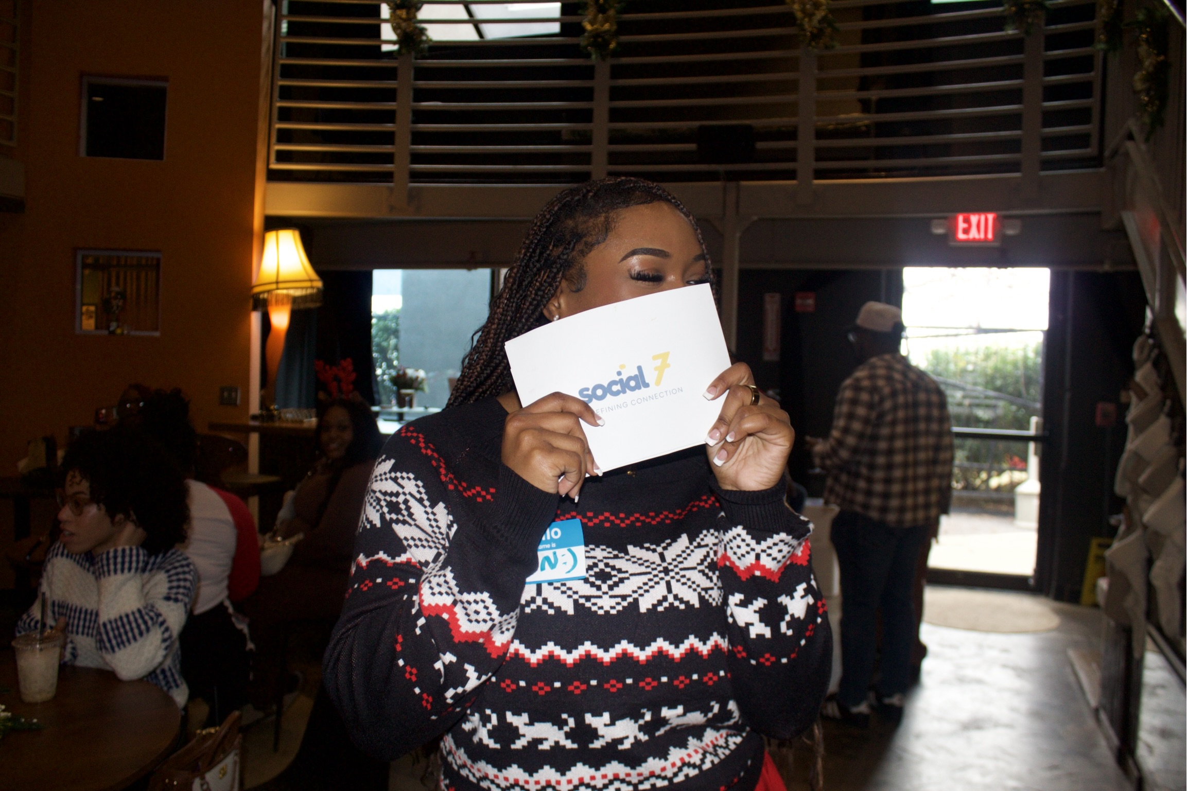 Person holding a card in a restaurant, with other people and a balcony in the background.
