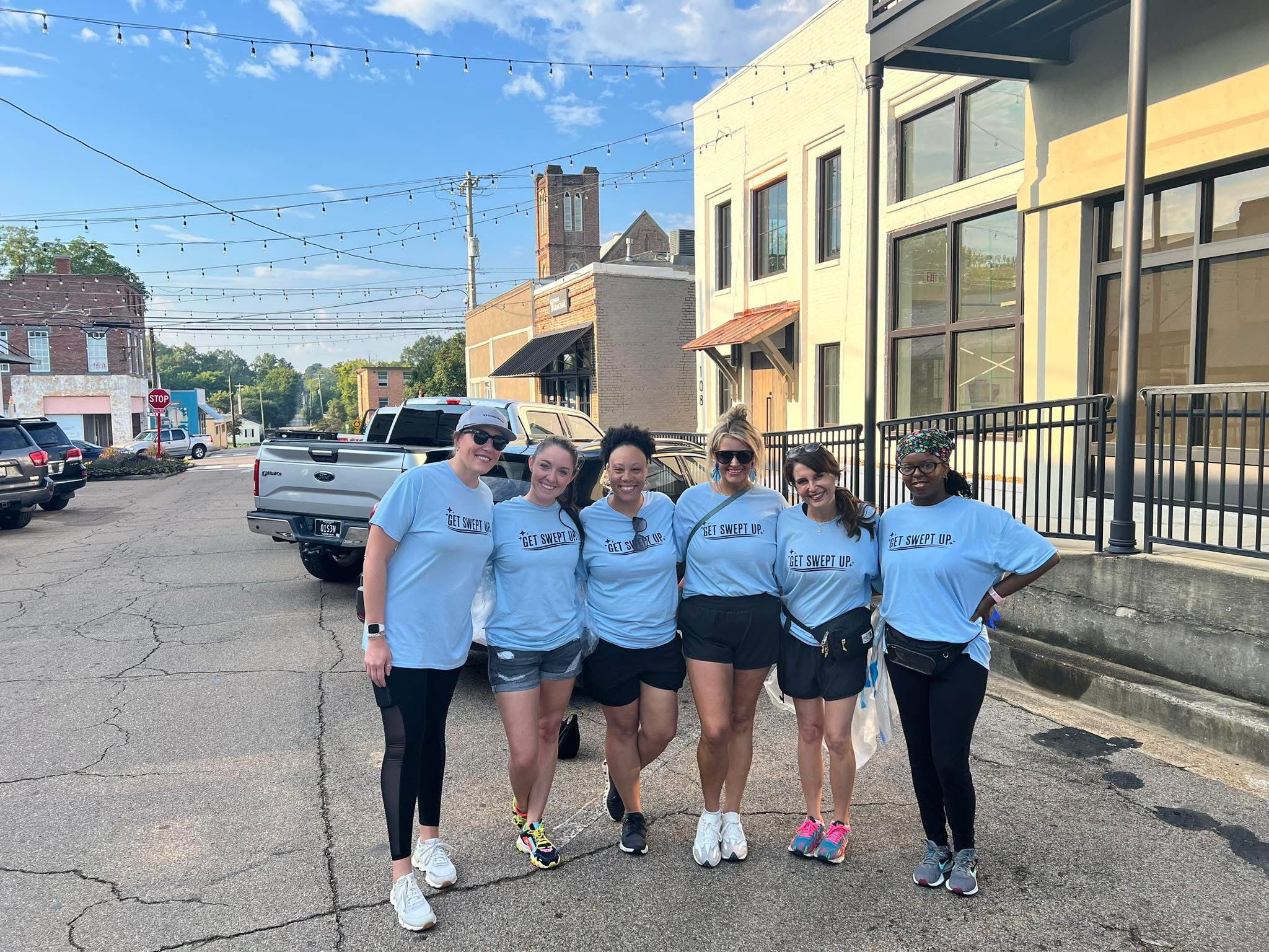 Group of six people standing outdoors on a street, smiling, wearing matching light blue shirts, with buildings and parked cars in background.