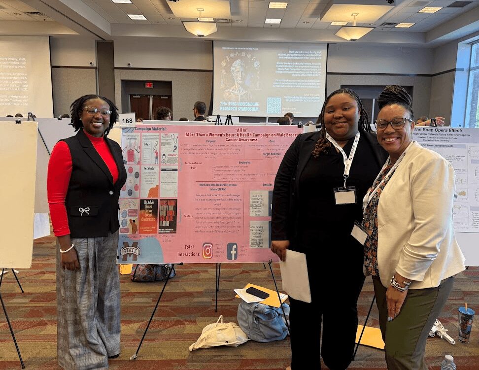 Three women standing in front of a research poster at a conference, smiling, in a large room with presentation screens.
