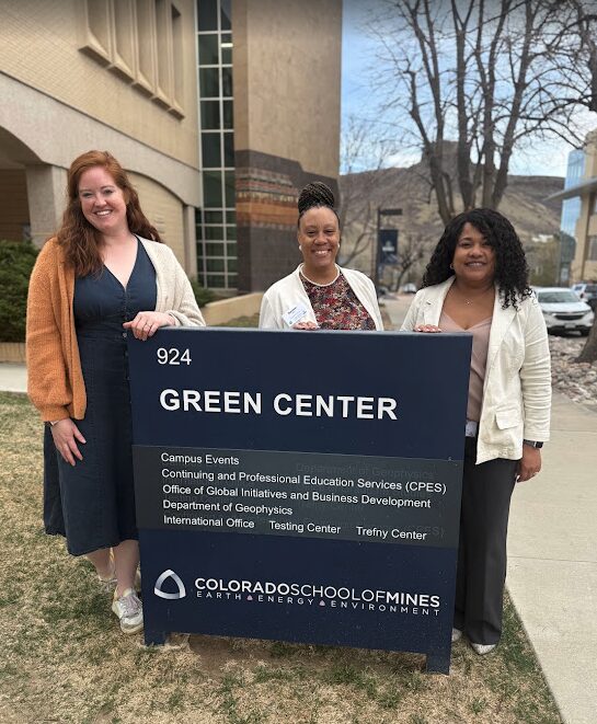 Three women standing outdoors behind a large sign for Green Center, smiling, with trees and buildings in the background.