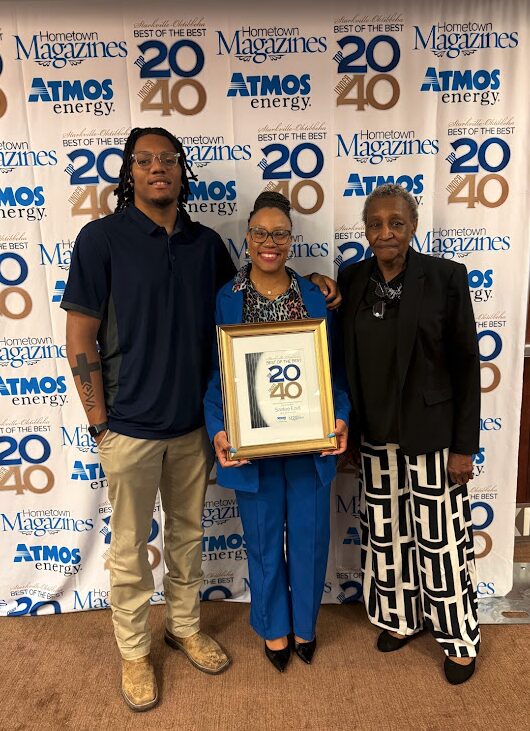 Three people standing in front of a backdrop with logos and text, holding a framed award, smiling.