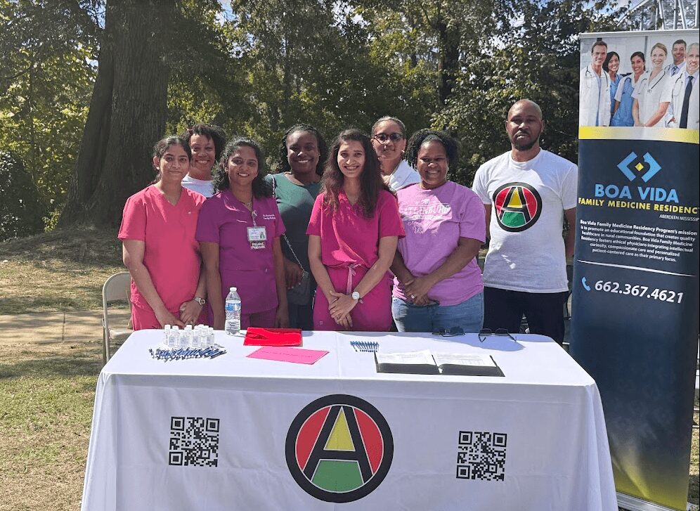 Group of seven people standing outdoors behind a table with a logo, trees in background, sunny day.