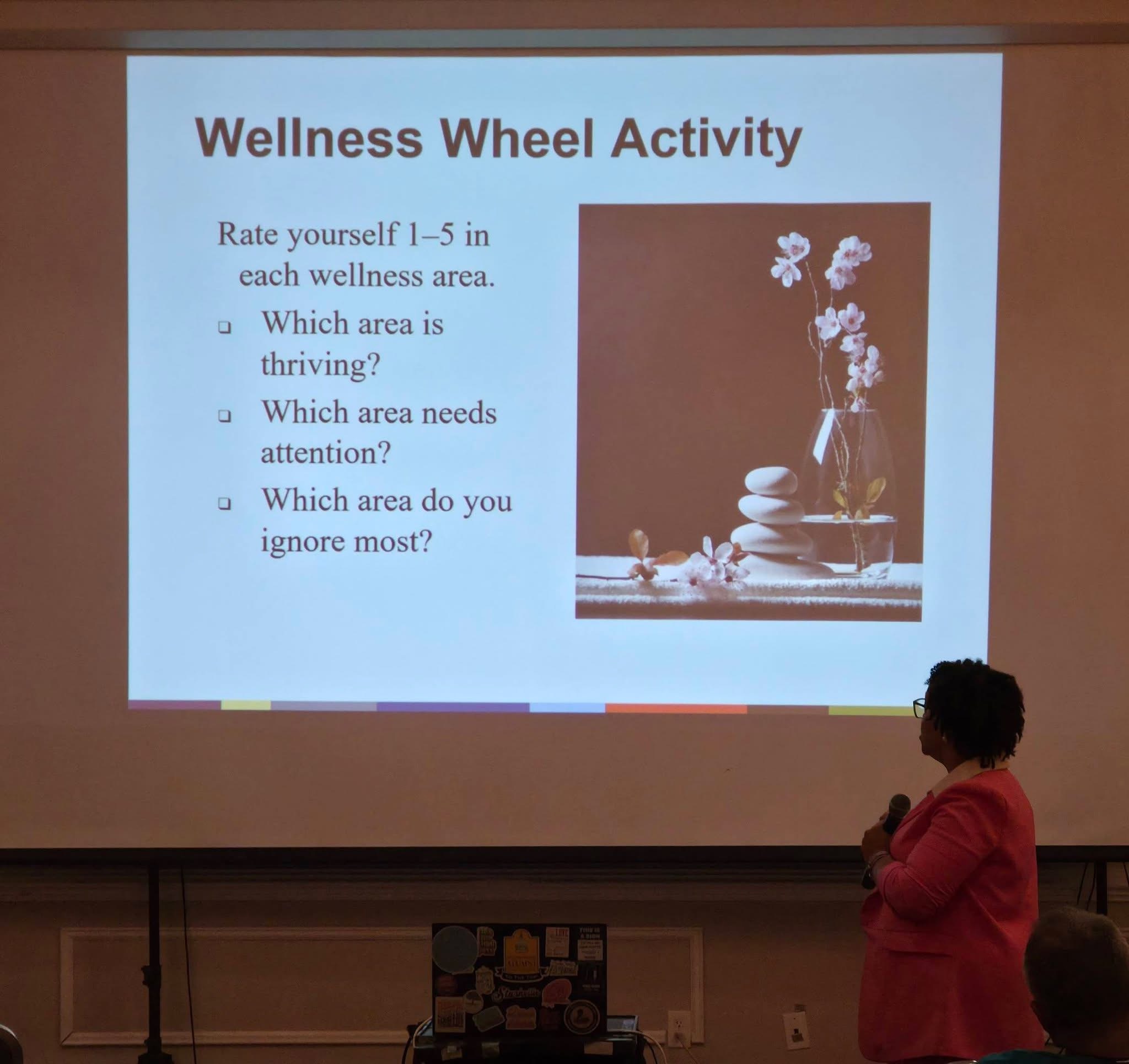 A woman in a pink jacket presents a slide titled 'Wellness Wheel Activity' with questions about wellness areas, on a large screen.