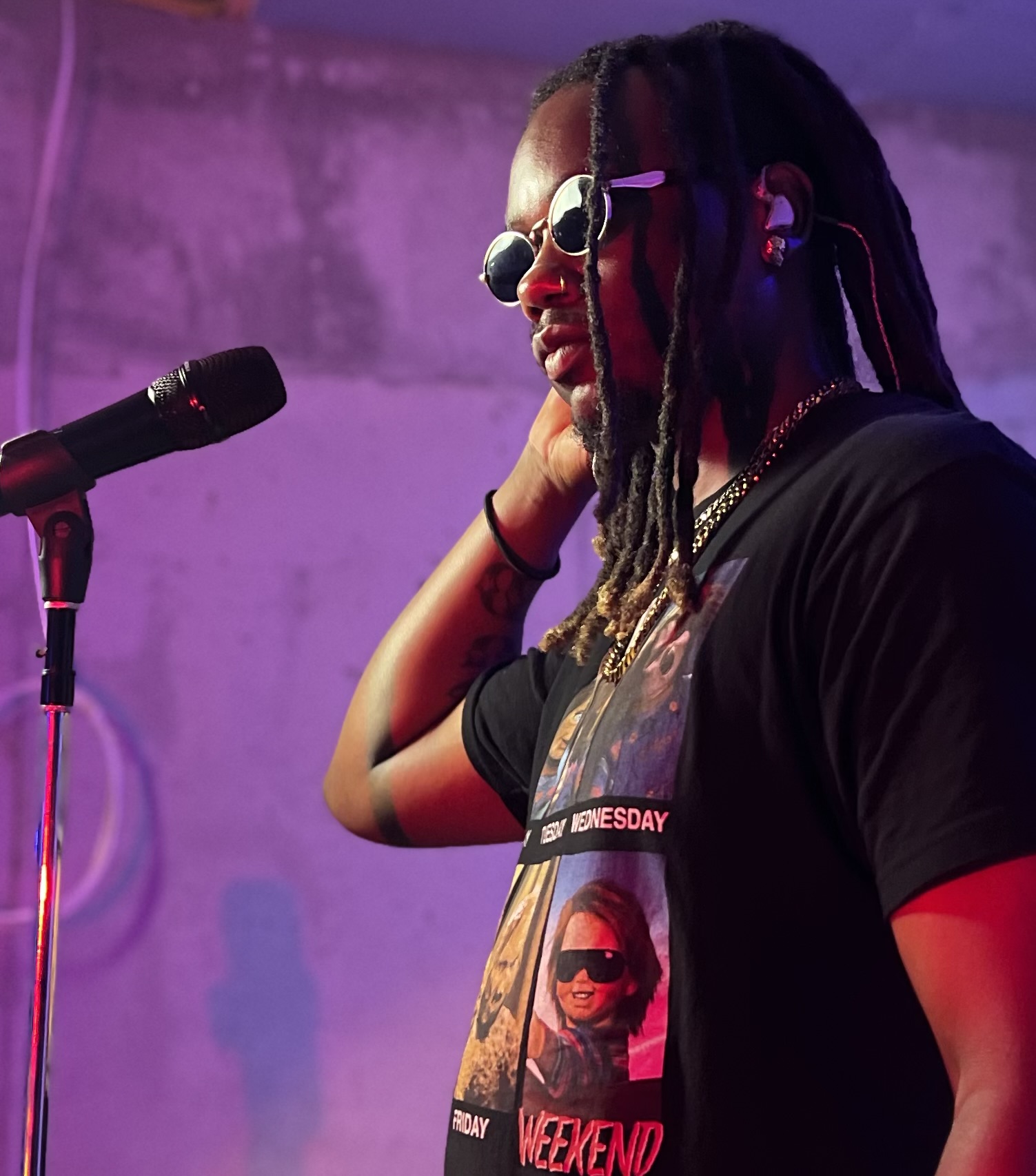 Person with dreadlocks wearing sunglasses and a black T-shirt, standing near a microphone, in a purple-lit room.