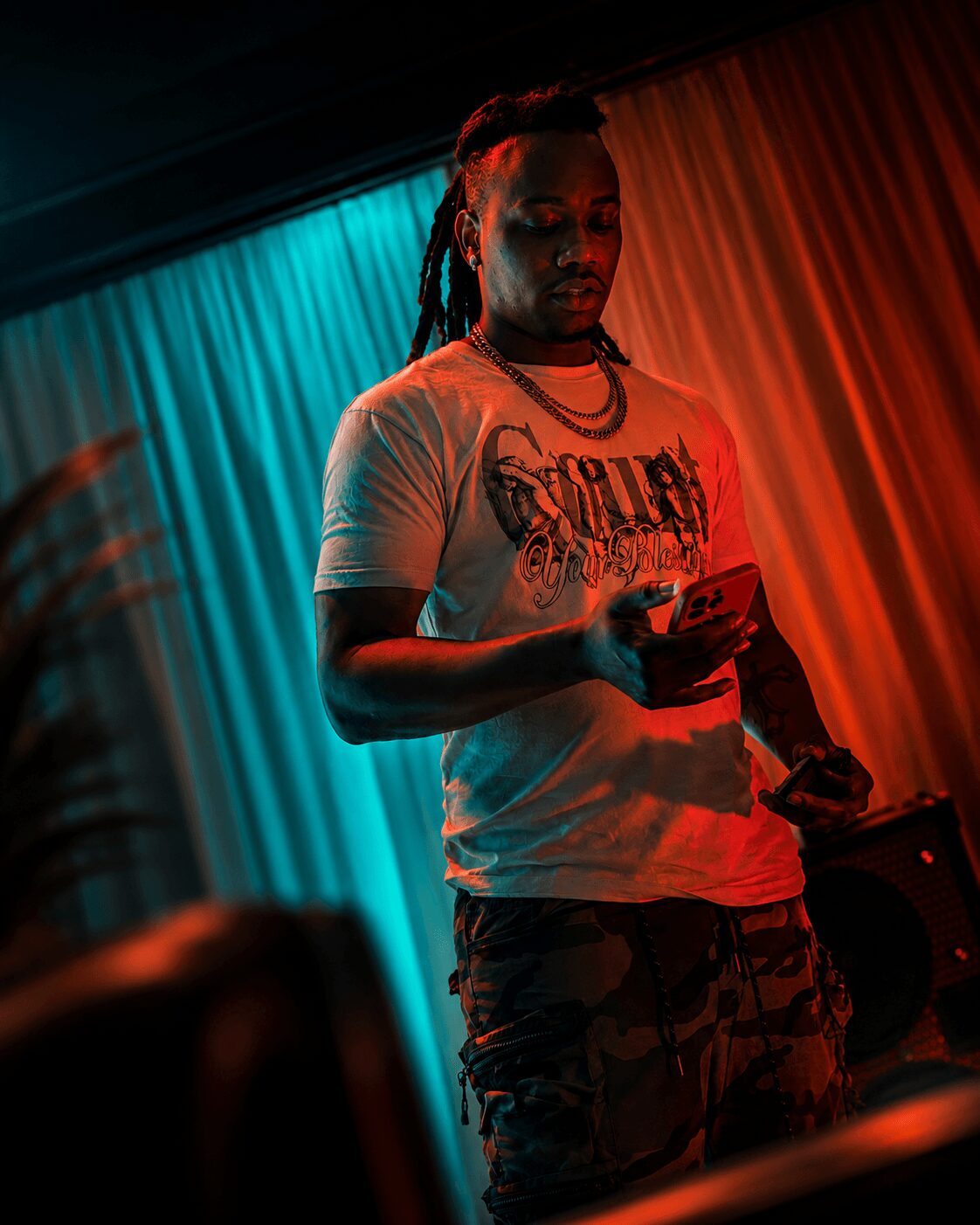 Person with dreadlocks looking at a phone, standing in front of colorful curtains, illuminated by red and blue lights.