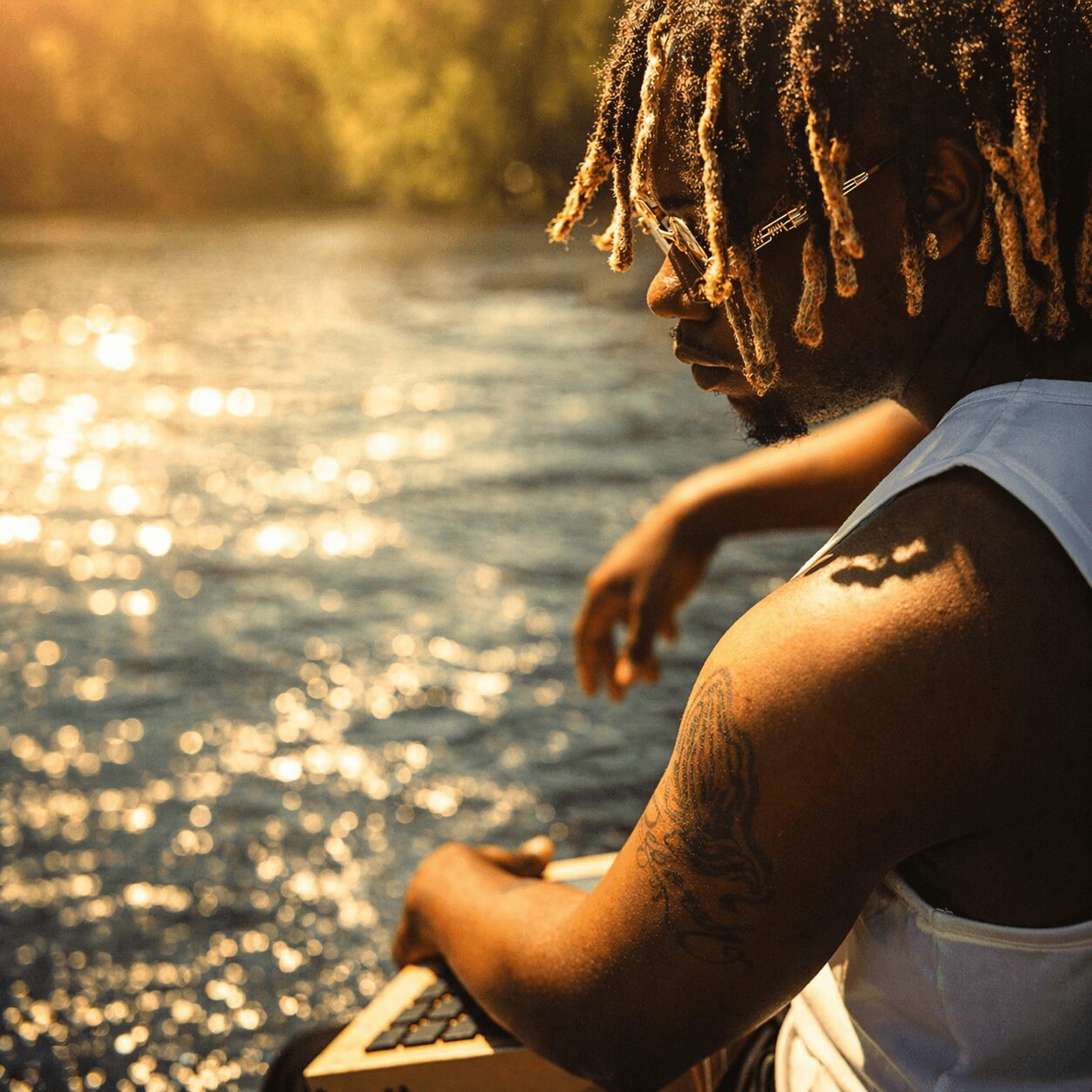 Person with dreadlocks and tattoos sitting by a river during sunset, arm resting on knee, water shimmering in background.