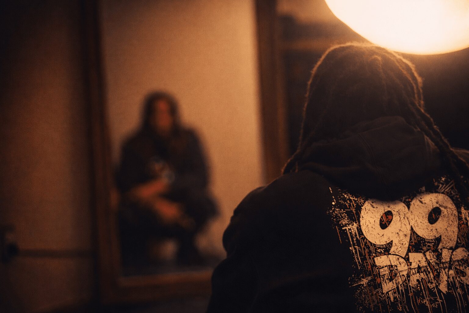 Person with dreadlocks facing away, looking at a mirror reflecting a person sitting on the floor, warm lighting.