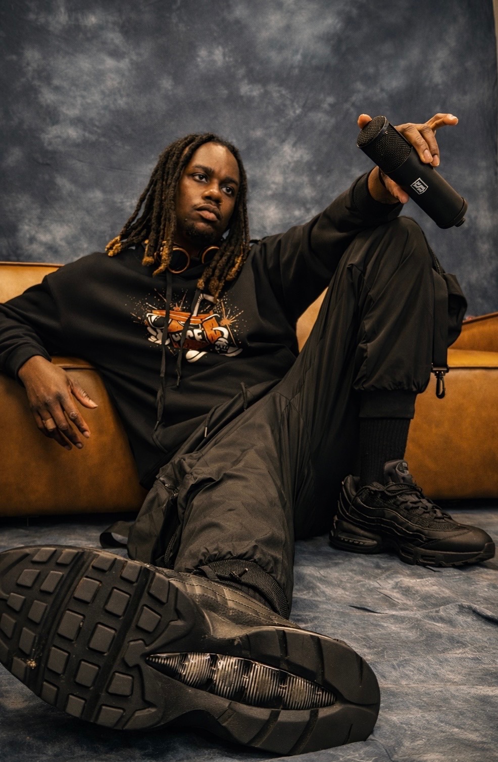 Person with dreadlocks sitting on a couch, holding a microphone, wearing black clothing and boots, against a dark background.