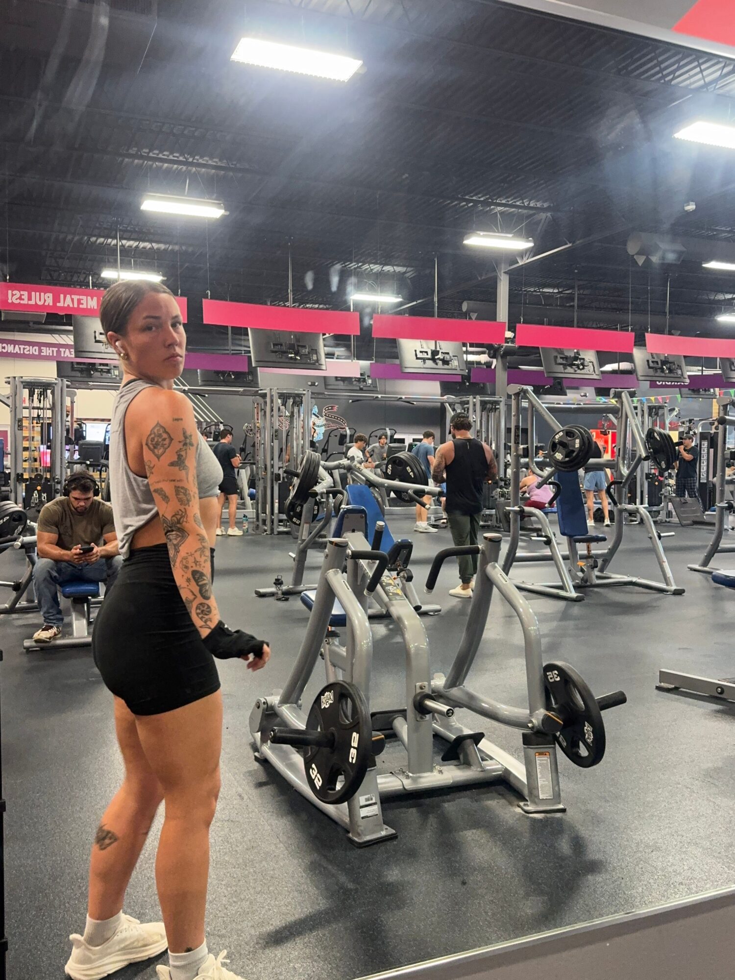A woman with tattoos on her arms and leg, wearing black shorts and a sports bra, stands in a gym near exercise equipment.