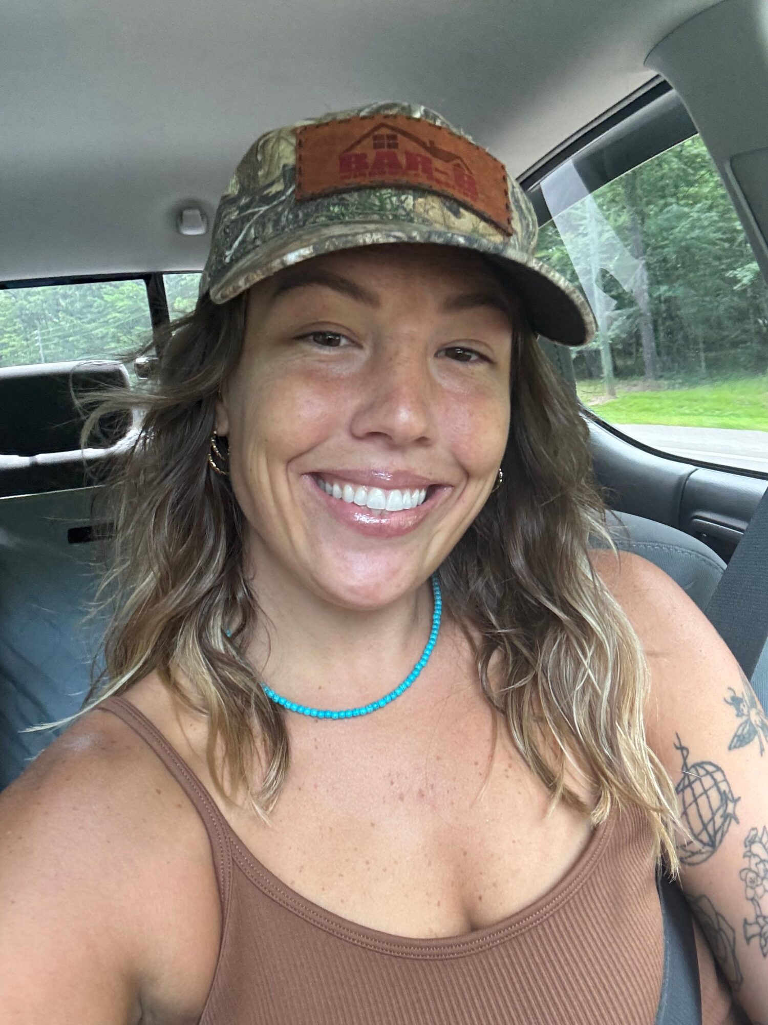 Woman smiling, wearing a hat and a brown tank top, sitting in a vehicle with a window showing greenery outside.