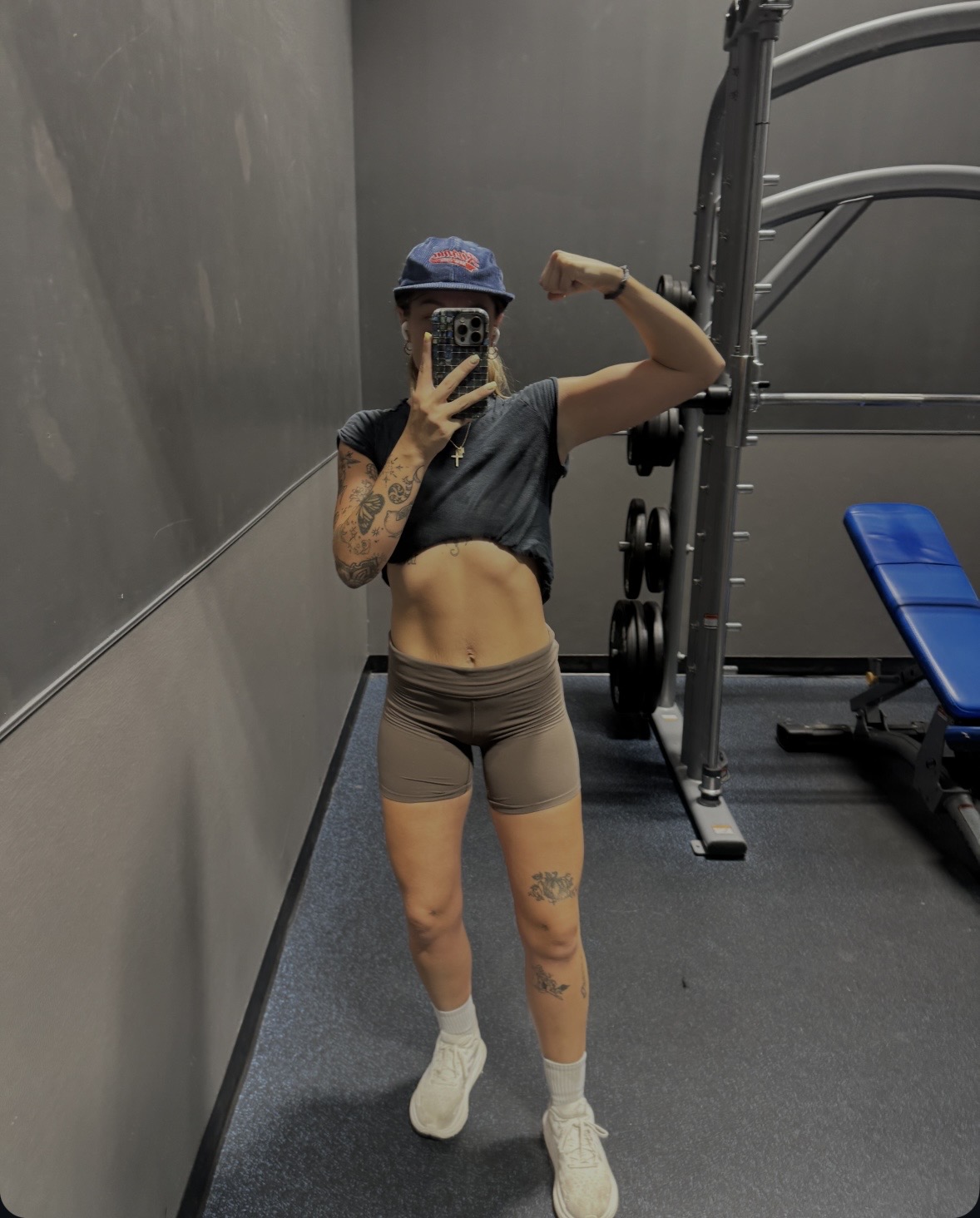 Person in gym taking mirror selfie, flexing arm, wearing cap, crop top, shorts, and sneakers, with gym equipment in background.