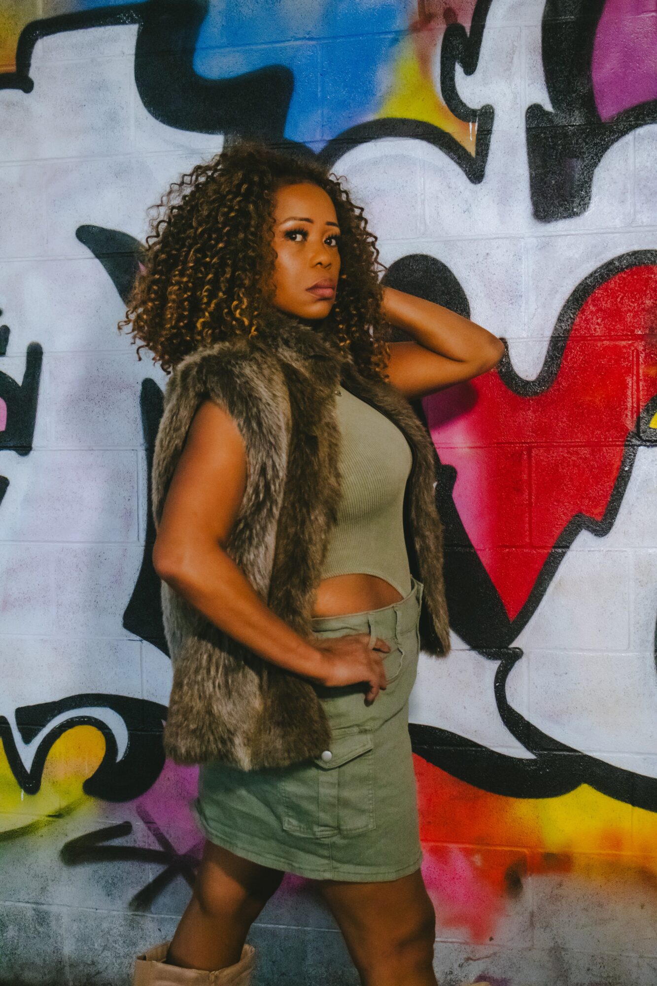 Woman with curly hair standing against graffiti wall, wearing a fur vest and olive green outfit.