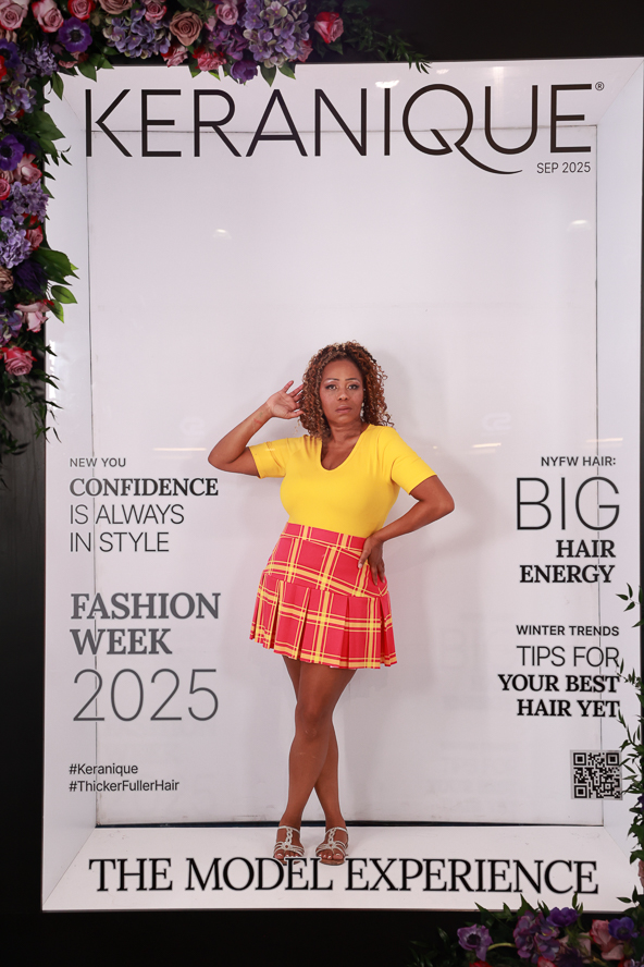 Woman in yellow top and red plaid skirt standing in front of a white backdrop with text, saluting with her right hand.