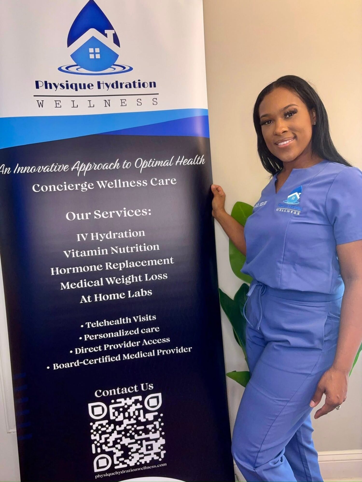 Woman in blue scrubs standing next to a wellness center banner, smiling, holding the banner with her right hand.