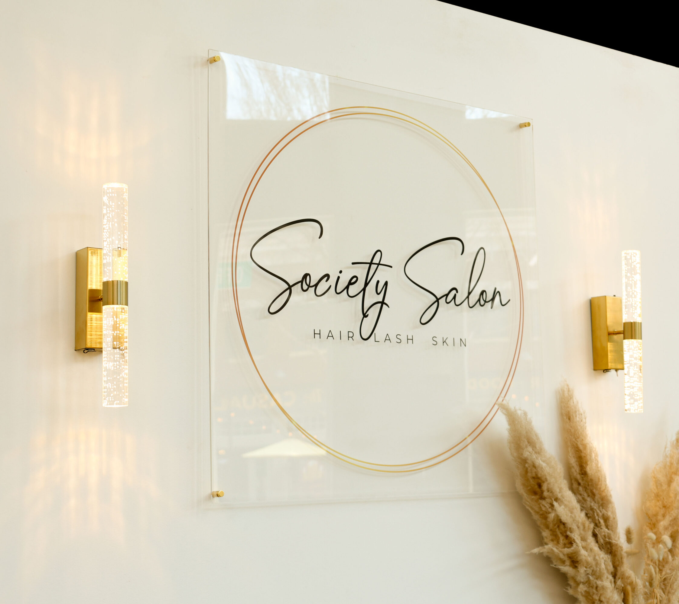 Sign reads 'Society Salon' with additional text below, mounted on a white wall with two gold wall sconces on each side.