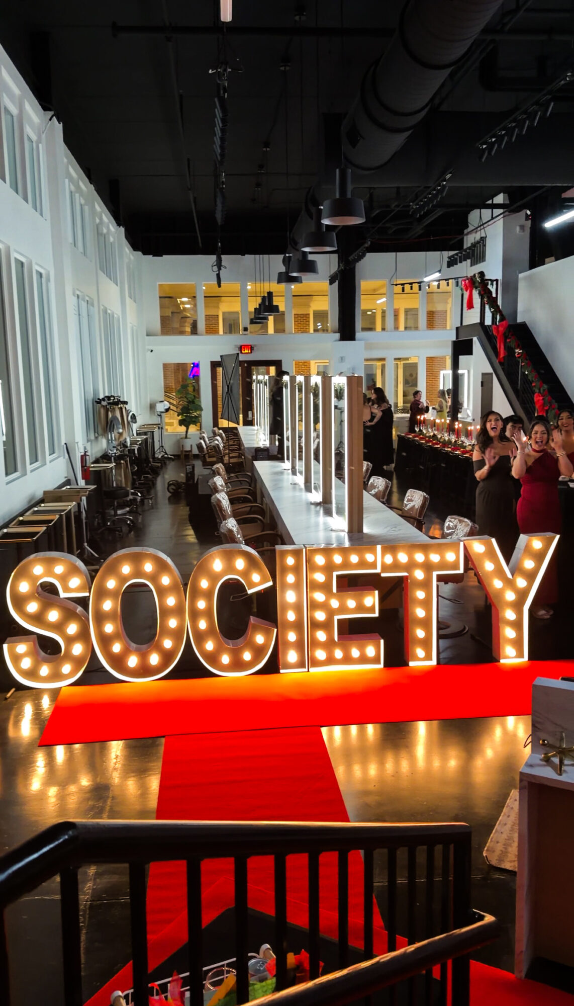 Interior of a social event with people, tables, and bright lighting, with a large illuminated sign reading 'SOCIETY' in the foreground.