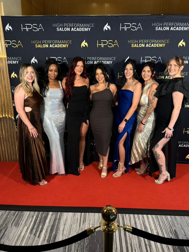 Seven women in formal dresses stand on a red carpet in front of a black backdrop with logos, smiling for a photo.