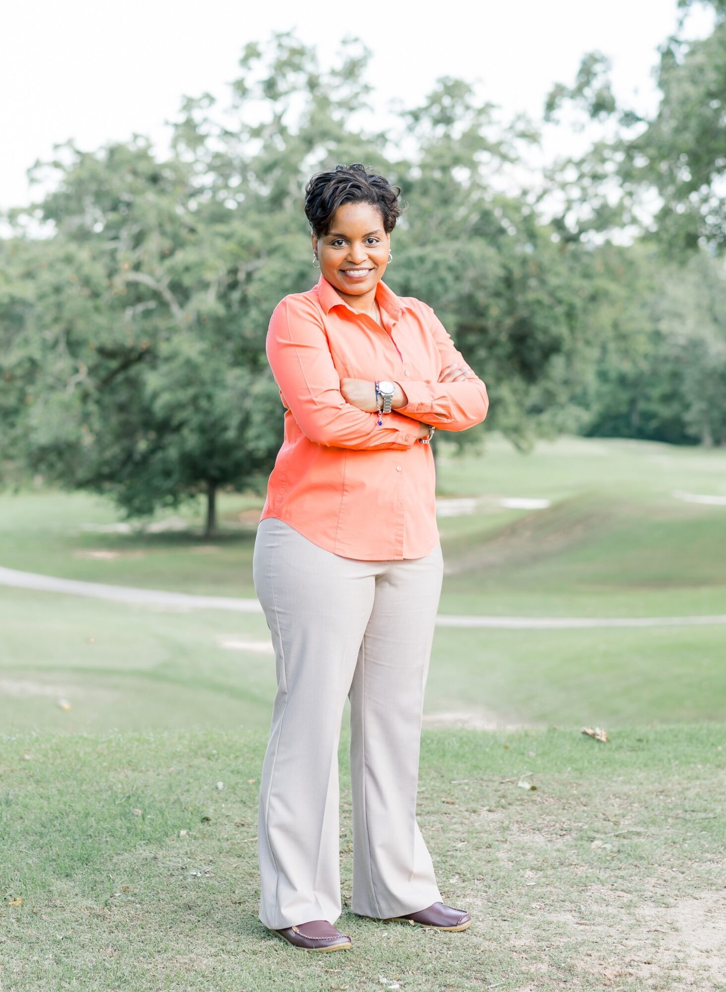 Woman standing outdoors with crossed arms, smiling, in a park with trees and grass.
