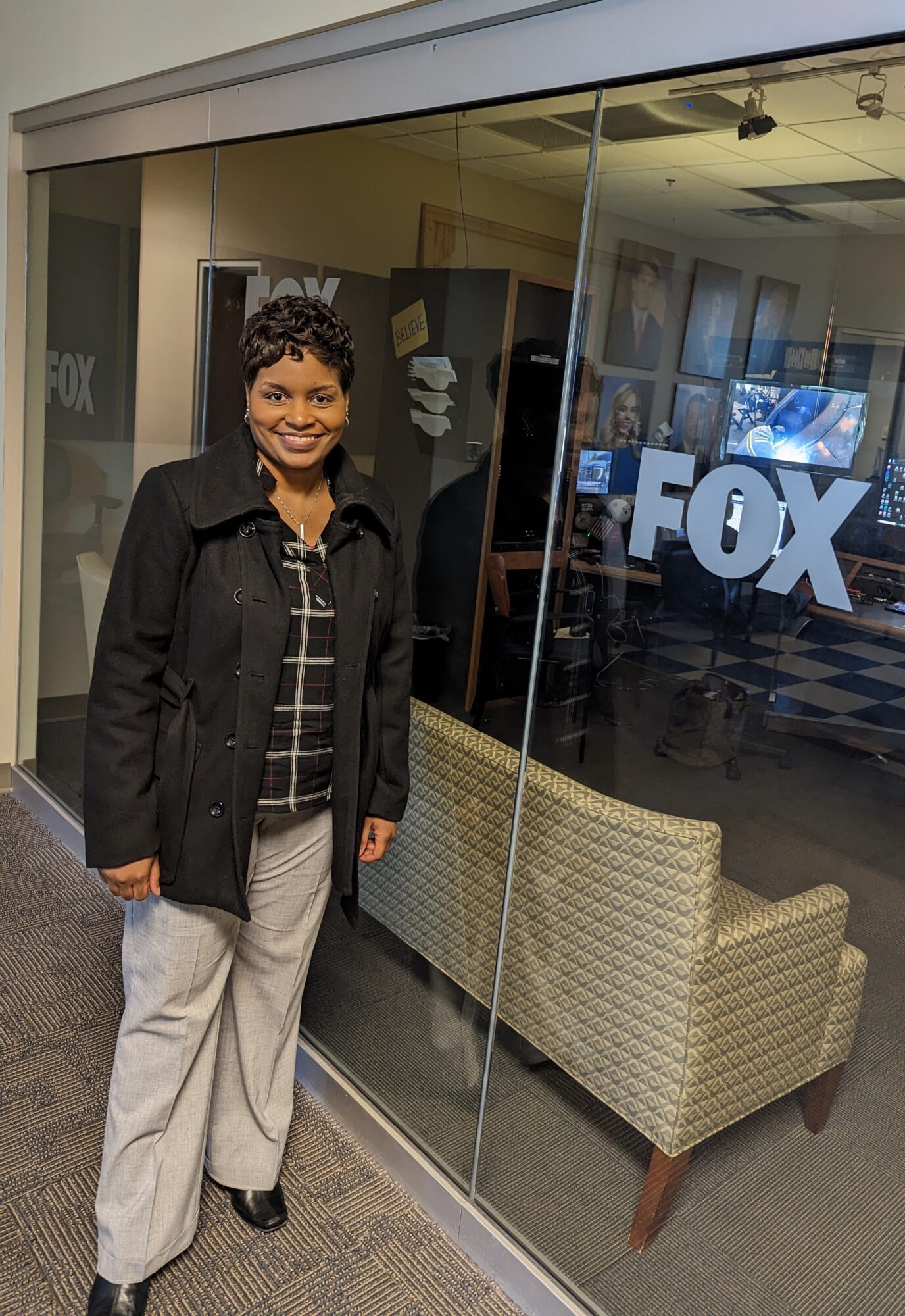 Woman standing outside glass window with FOX logo, smiling, wearing a black jacket and light-colored pants.