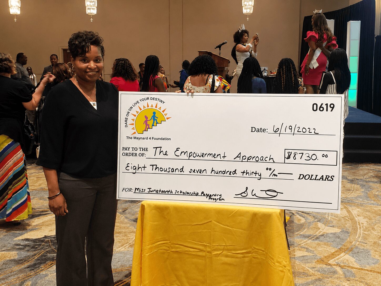 Woman standing next to a large check at an indoor event, with people in the background.
