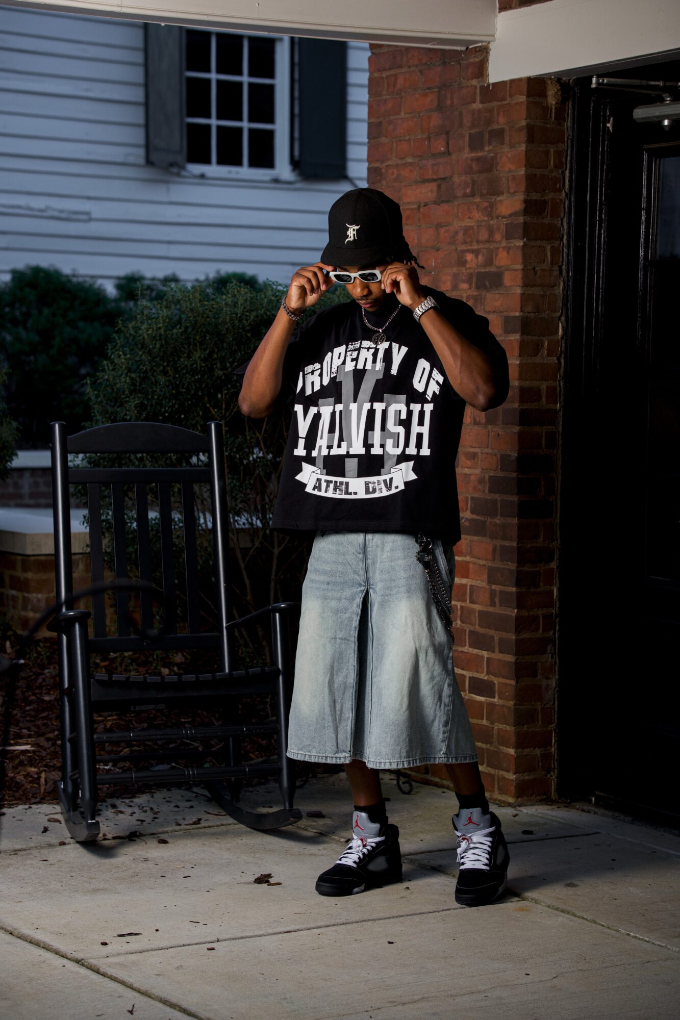 Young person standing outside, wearing a black cap, sunglasses, oversized black T-shirt, and baggy shorts, holding sunglasses.