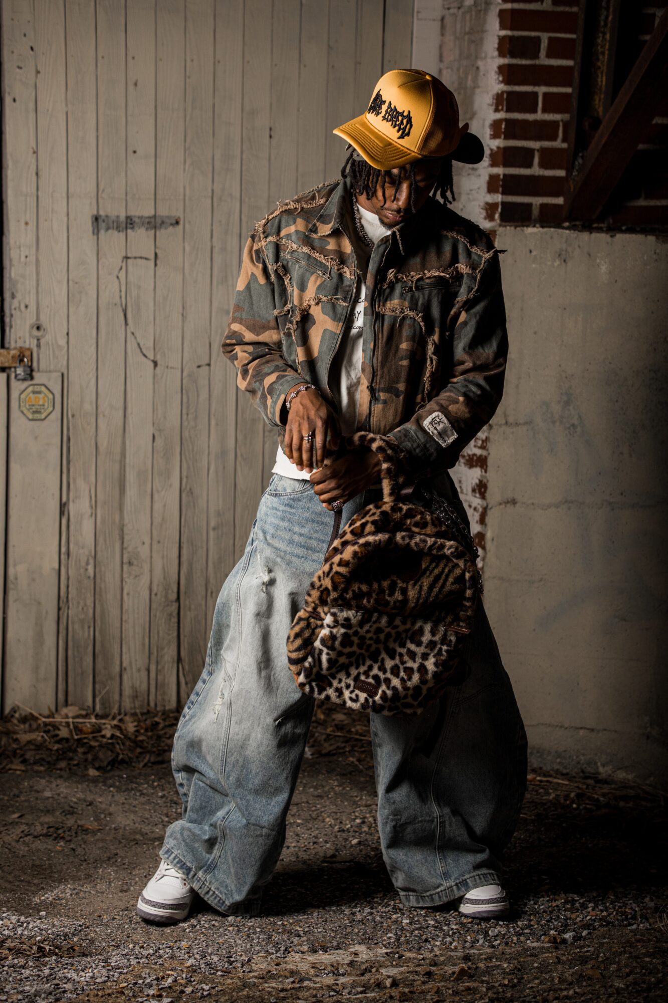 Person wearing a yellow cap, camouflage jacket, and baggy jeans, holding a leopard-print bag, standing outdoors against a wall.
