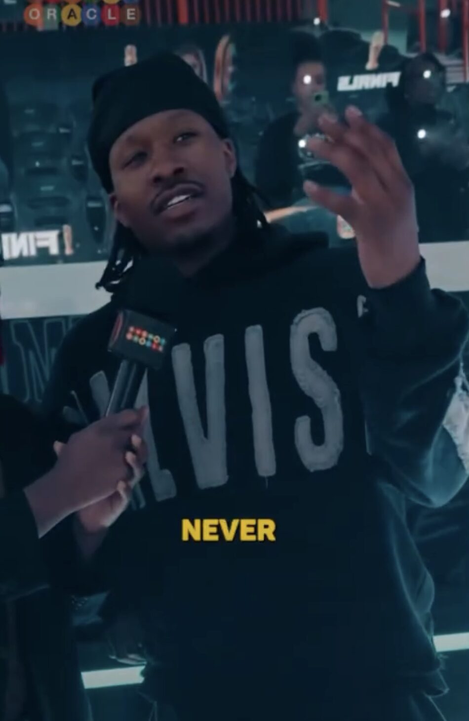 Person wearing a black hoodie with large gray letters, speaking into a microphone, with a crowd and lights in the background.