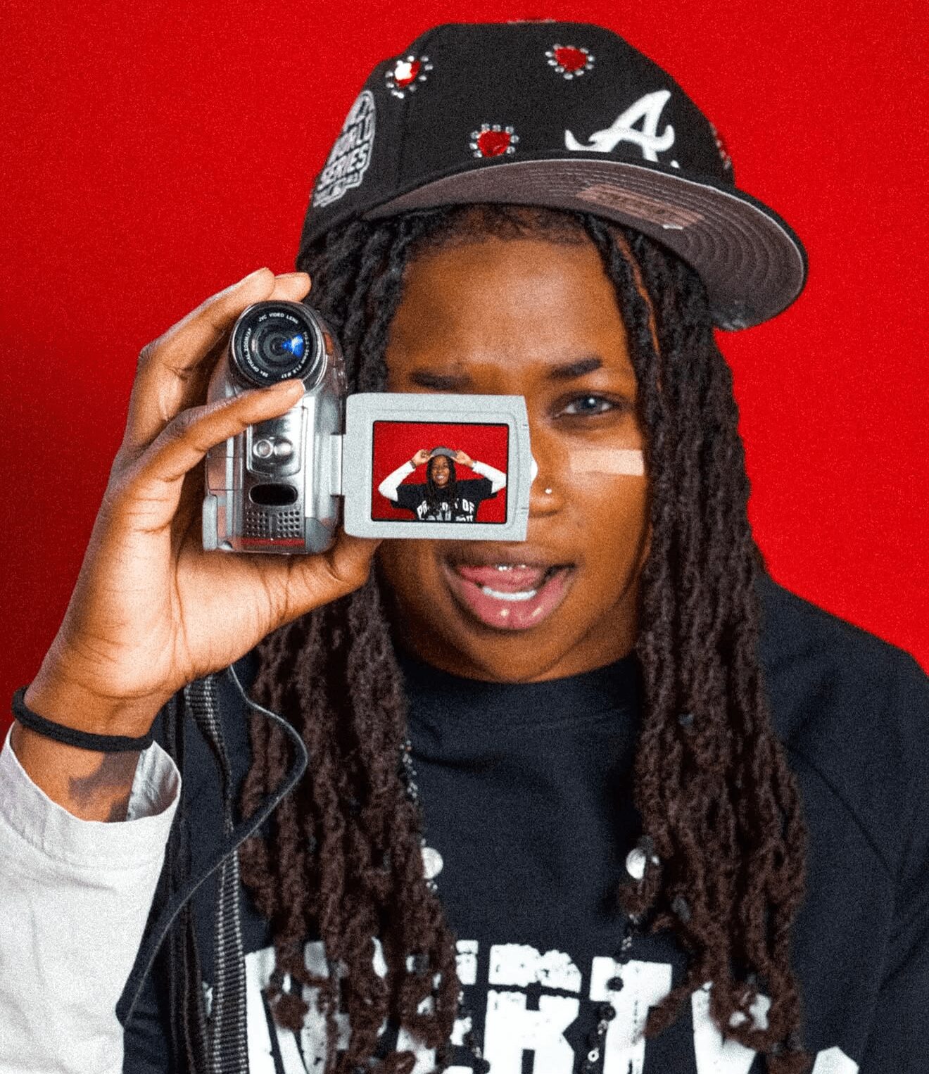 Person with dreadlocks holding a camera, wearing a black cap and black shirt, against a red background.